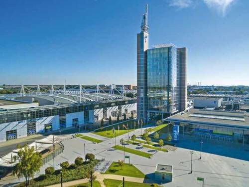 Luftaufnahme des Messegeländes Hannover mit Glasbürogebäude, Funkturm, Messehallen und weitem Vorplatz bei sonnigem Wetter.