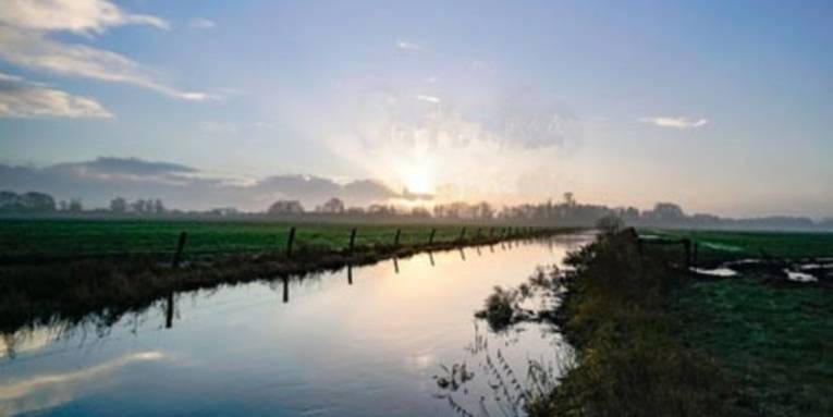 Sonnenaufgang über einer weiten Wiesenlandschaft mit einem ruhigen Wassergraben, der den Himmel reflektiert.